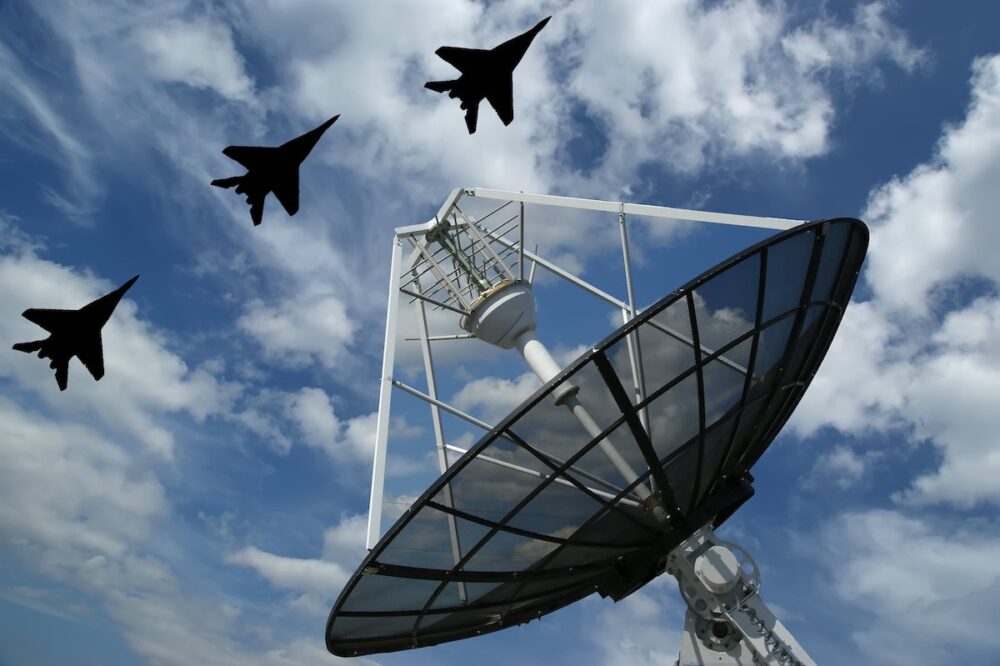 A radar dish points up toward three military jets flying overhead.