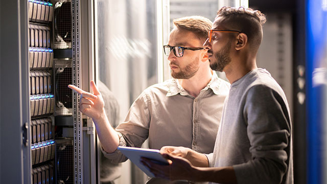 Team of young multi-ethnic specialists in glasses standing at server and using tablet while managing network server together in data center